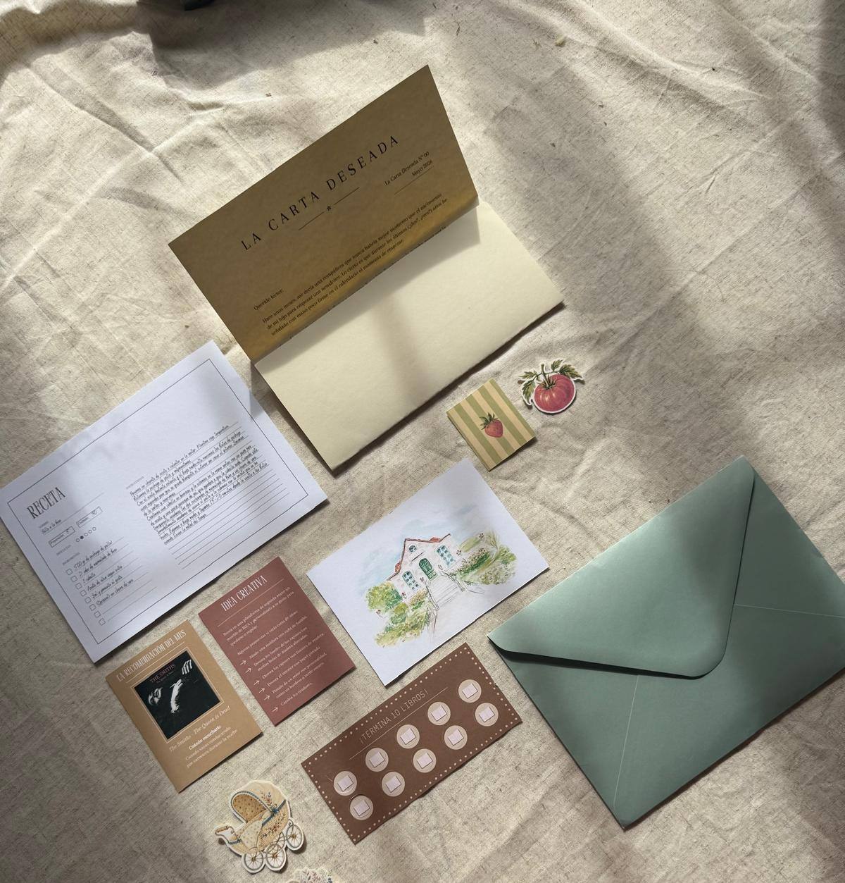 Contenido de La Carta Deseada sobre una tela: carta, receta, ilustración, pegatinas y un sobre verde.